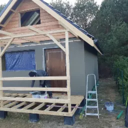 Budowa małego, murowanego domu w stanie surowym otwartym, z widocznym ociepleniem ścian zewnętrznych i konstrukcją drewnianą werandy. Pracownik nakłada zaprawę na elewację, obok drabina i wiadro...