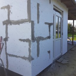 Usługi ogólnobudowlane - Ocieplanie elewacji budynku styropianem z widocznymi paskami kleju mocującego, przygotowanie do nałożenia siatki i tynku.