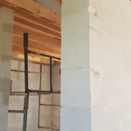 Wnętrze w trakcie budowy: ściany z białych bloczków, widoczne drewniane krokwie dachu i metalowe rusztowanie.