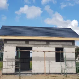 Niedokończony budynek z białych bloczków z zamontowanym ciemnym dachem, widoczne rusztowania i błękitne niebo z chmurami w tle.