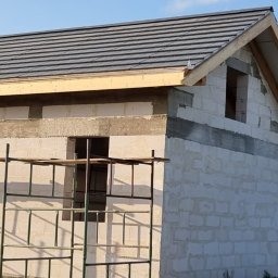 Usługi ogólnobudowlane - Surowy stan domu z białych bloczków, z szarą dachówką, drewnianą konstrukcją dachu, otworami okiennymi i rusztowaniem, na tle zielonego terenu i błękitnego nieba.