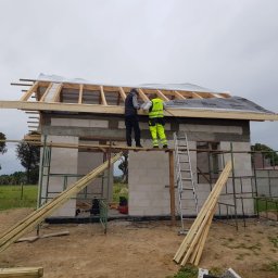 Usługi ogólnobudowlane - Budowa domu z bloczków: dwóch robotników montuje konstrukcję dachu na niewykończonym budynku, widoczne rusztowania i materiały budowlane.