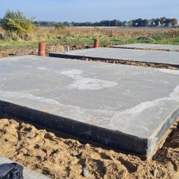Usługi ogólnobudowlane - Wylane płyty fundamentowe zabezpieczone czarną izolacją, widoczne rury kanalizacyjne wystające z ziemi, piaszczyste podłoże i krajobraz polny w tle.