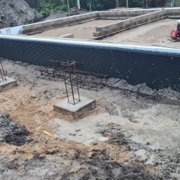 Usługi ogólnobudowlane - Etap budowy fundamentów: widoczne zbrojenia na betonowych podstawach, izolacja pionowa fundamentów z folii kubełkowej, częściowo zalany wykop, ubijarka w tle.