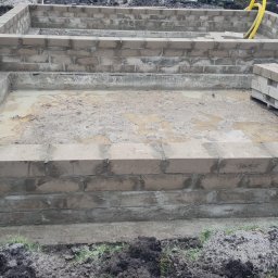 Usługi ogólnobudowlane - Wykopany fundament pod budynek z częściowo wymurowanymi ścianami z bloczków betonowych, widoczne stojące bloczki na palecie i żółta rura kanalizacyjna w ziemi.