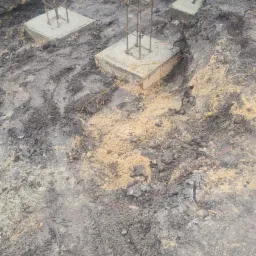 Wykopane fundamenty pod budynek z widocznymi betonowymi stopami i wystającymi zbrojeniami, otoczone mokrą ziemią i piaskiem.