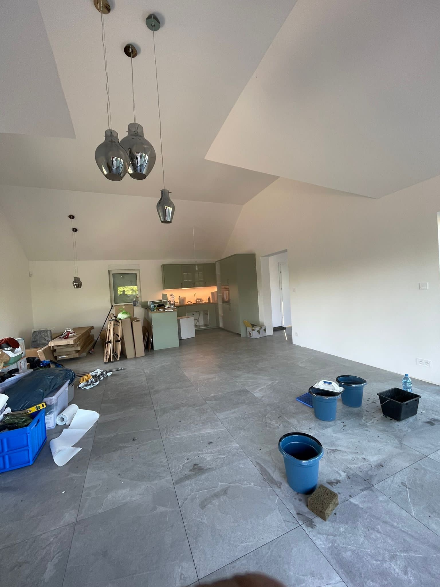 Wnętrze domu w trakcie prac wykończeniowych: widoczne wiadra z wodą i gąbka na szarych płytkach podłogowych, świeżo pomalowane białe ściany, w tle zabudowa kuchenna w odcieniach zieleni.