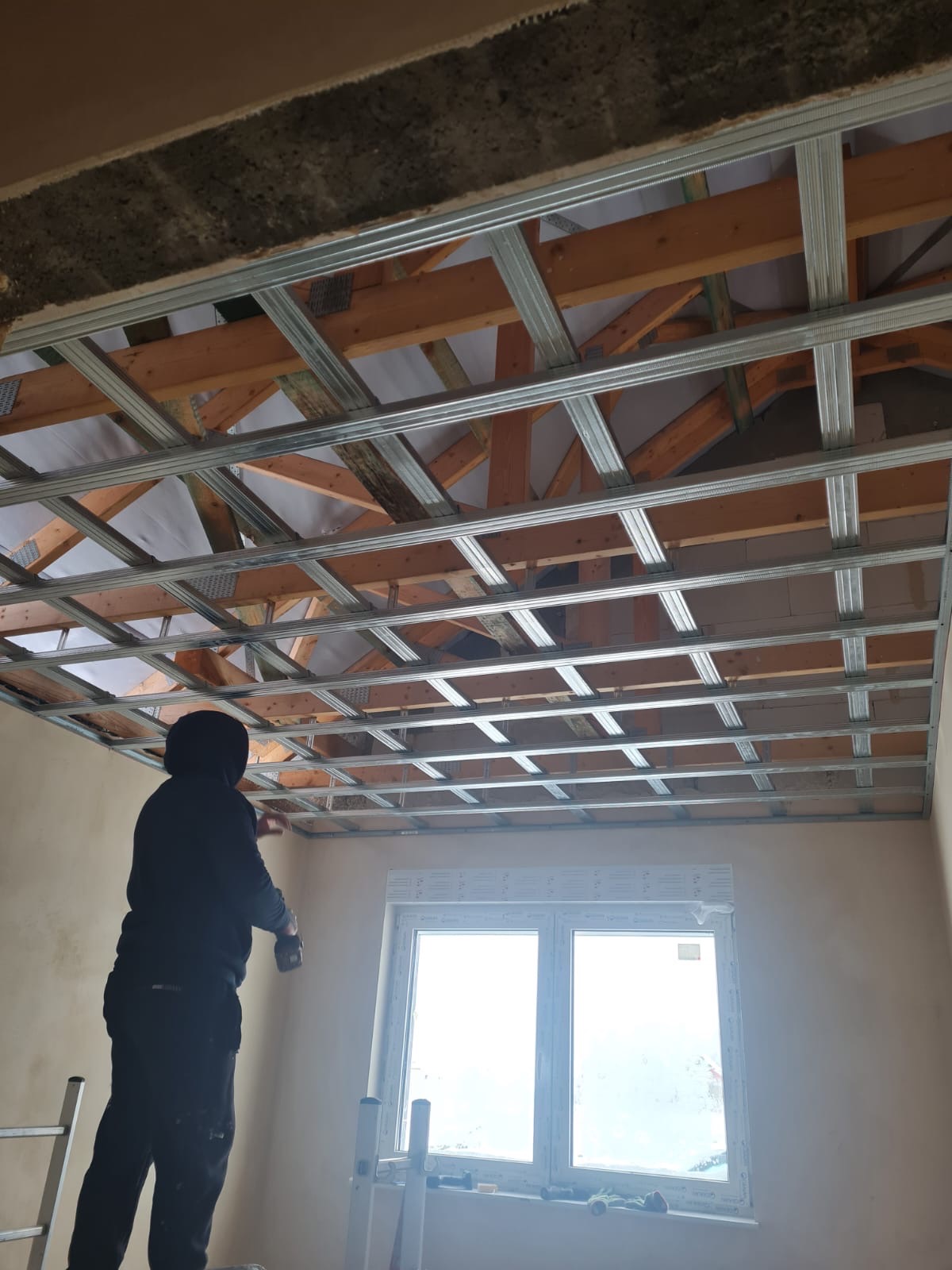 Montaż metalowej konstrukcji sufitu podwieszanego w Wolinie, widoczne drewniane elementy konstrukcyjne i biała izolacja na poddaszu.