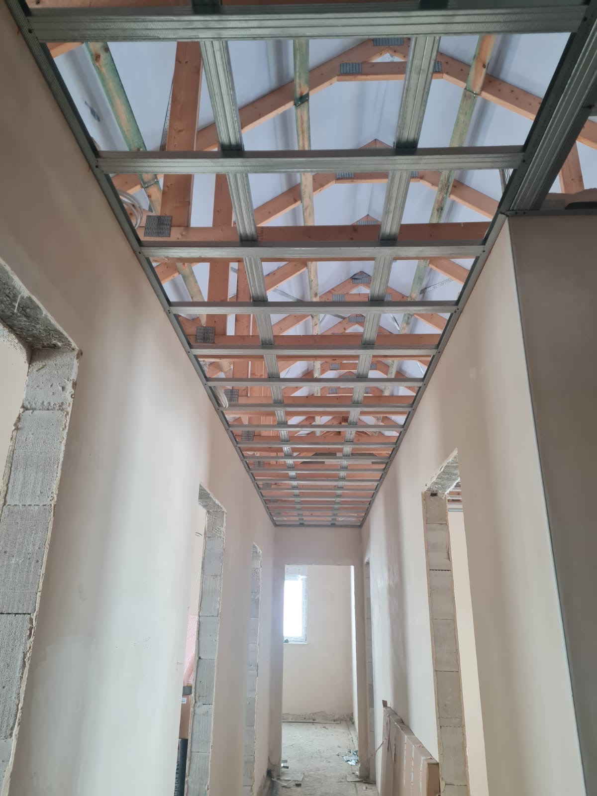 Wnętrze w trakcie wykańczania: widok na sufit z drewnianą konstrukcją i metalowymi profilami, ściany pokryte gładzią, otwory drzwiowe w trakcie obróbki.