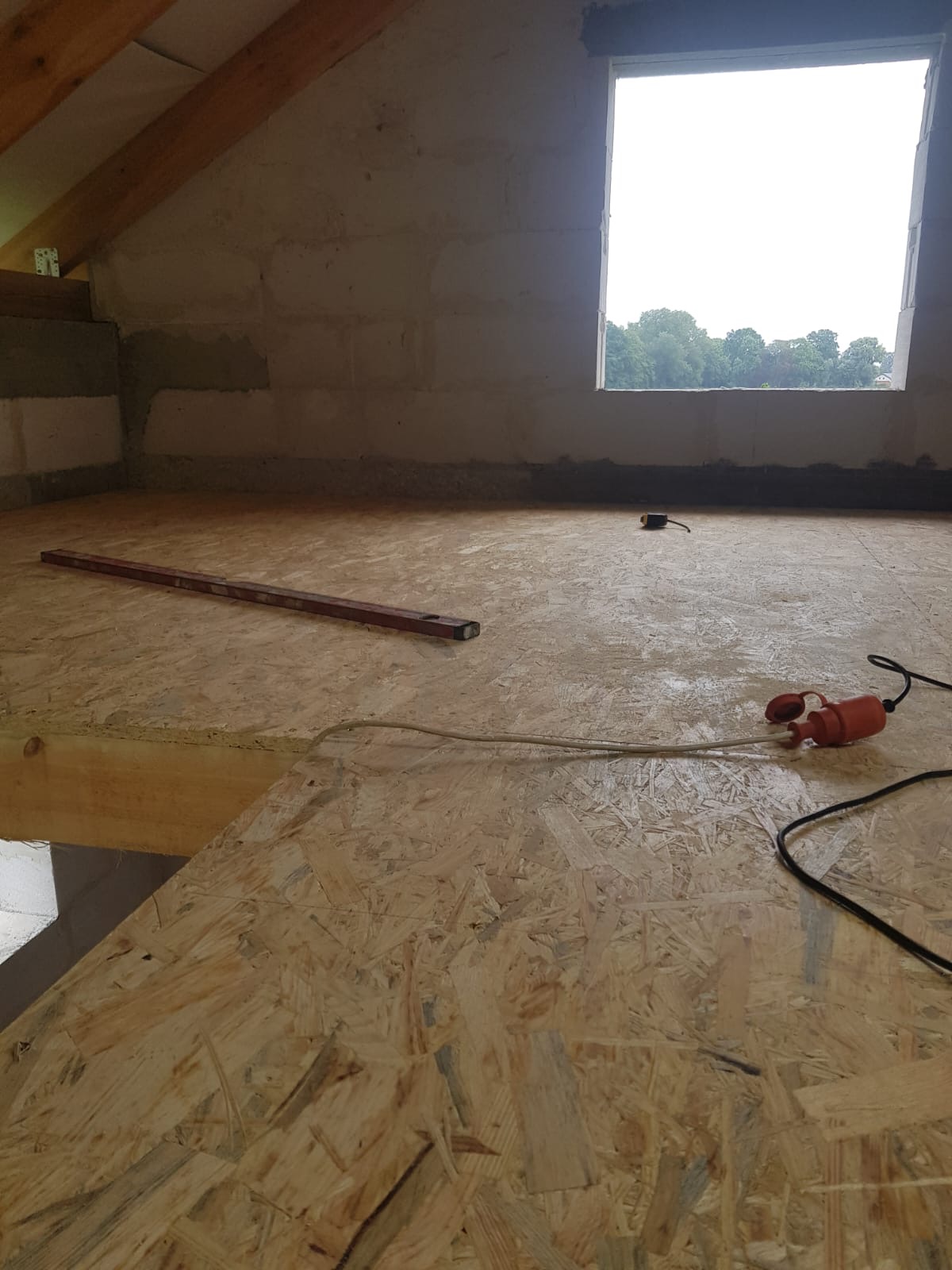Surowy stan poddasza z oknem, widoczne ściany z bloczków, drewniany strop z płyt OSB, poziomica i przedłużacz elektryczny leżące na podłodze.