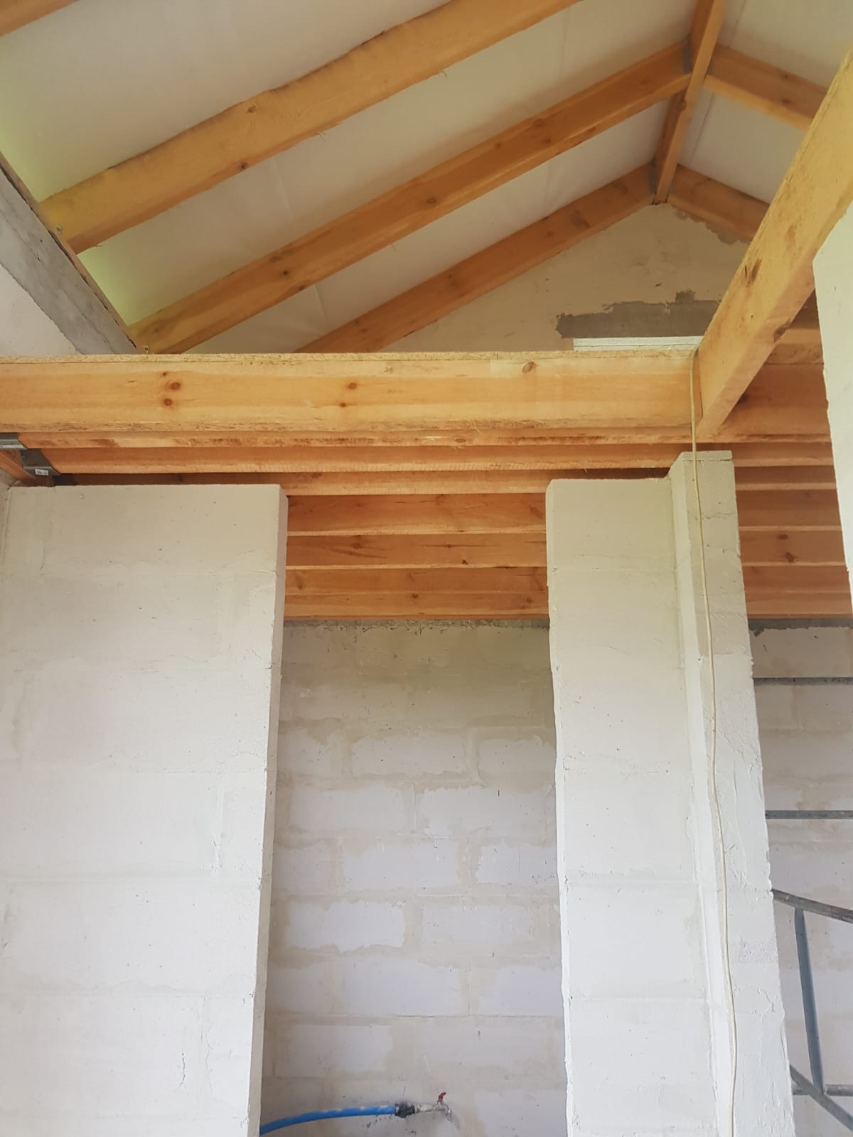 Wnętrze w trakcie budowy domu murowanego: ściany z bloczków, drewniana konstrukcja dachu, widoczne elementy instalacji wodnej przy podłodze.