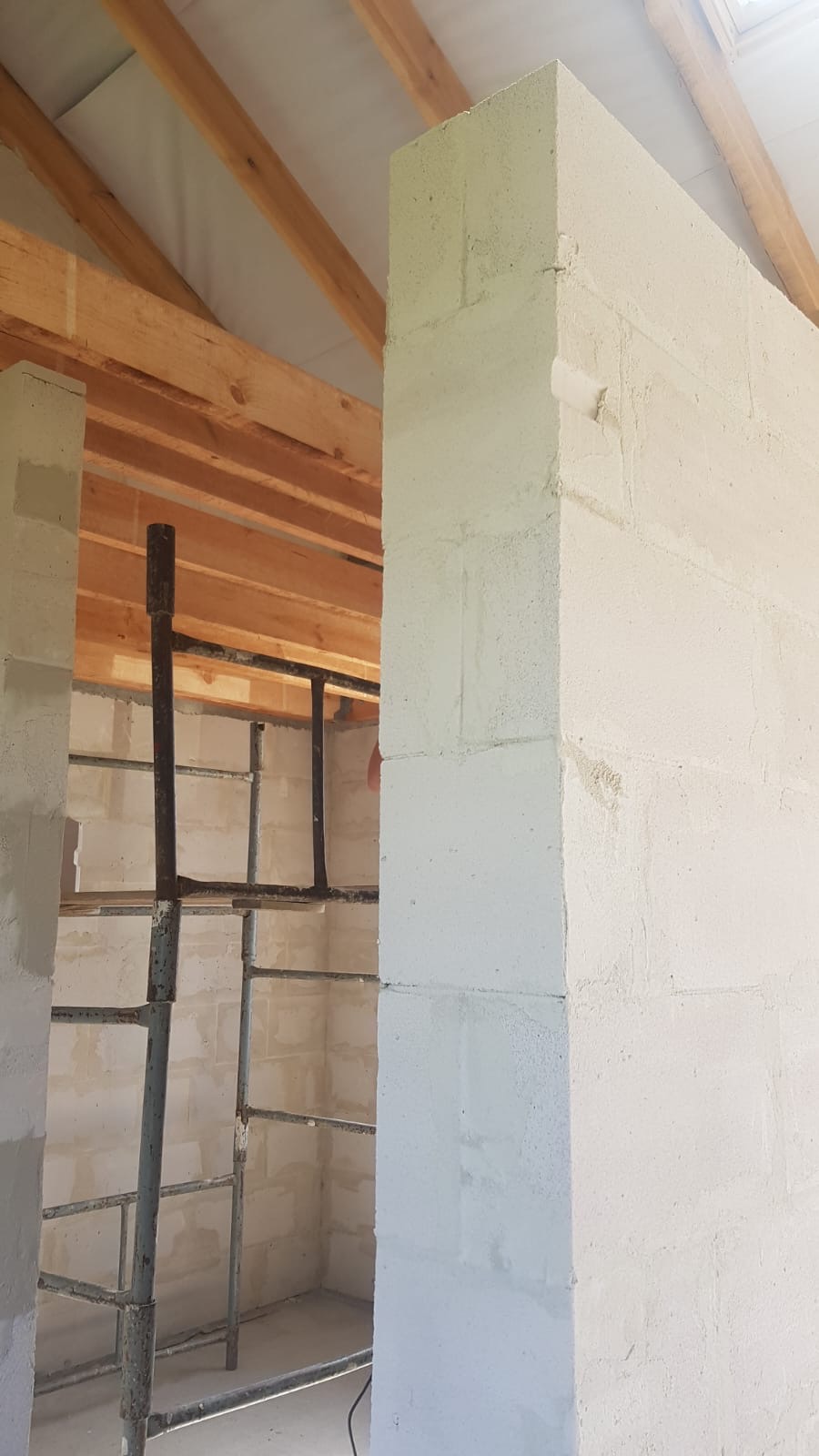 Wnętrze w trakcie budowy: ściany z białych bloczków, widoczne drewniane krokwie dachu i metalowe rusztowanie.