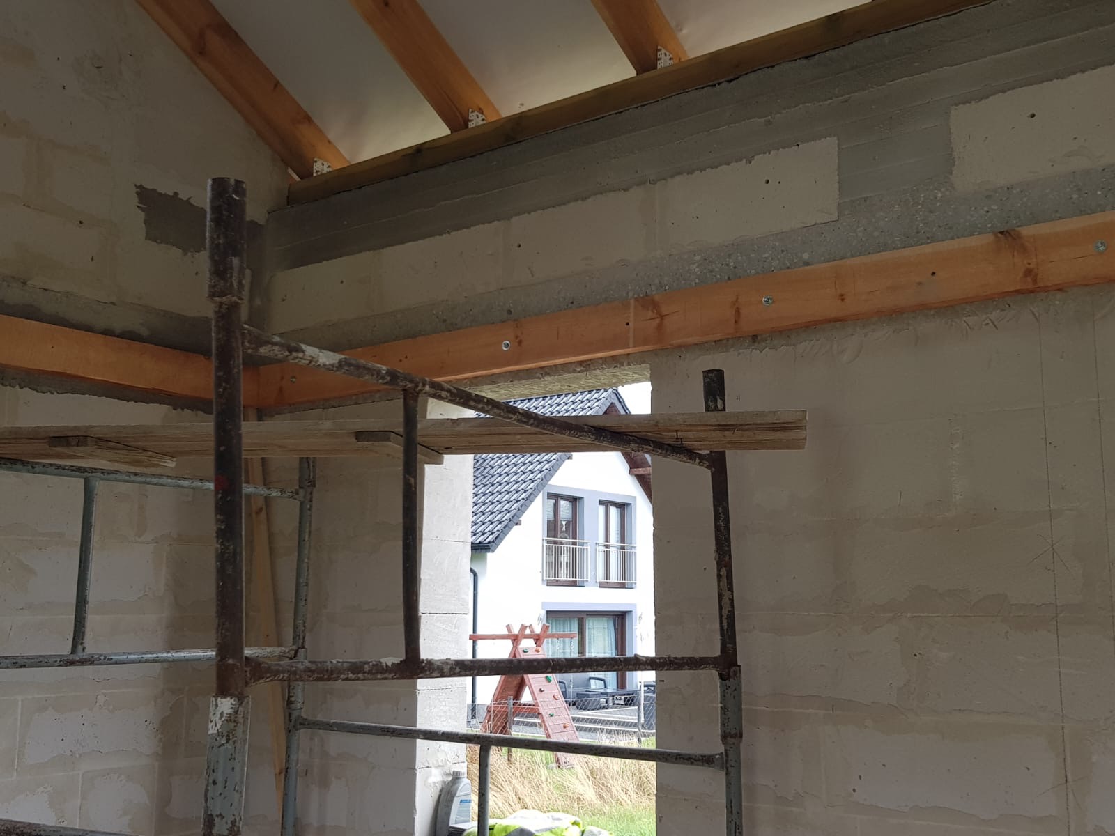 Wnętrze surowego domu w trakcie budowy, widoczne ściany z betonu komórkowego, drewniane belki stropowe, fragment rusztowania i widok na sąsiedni dom przez otwór okienny.