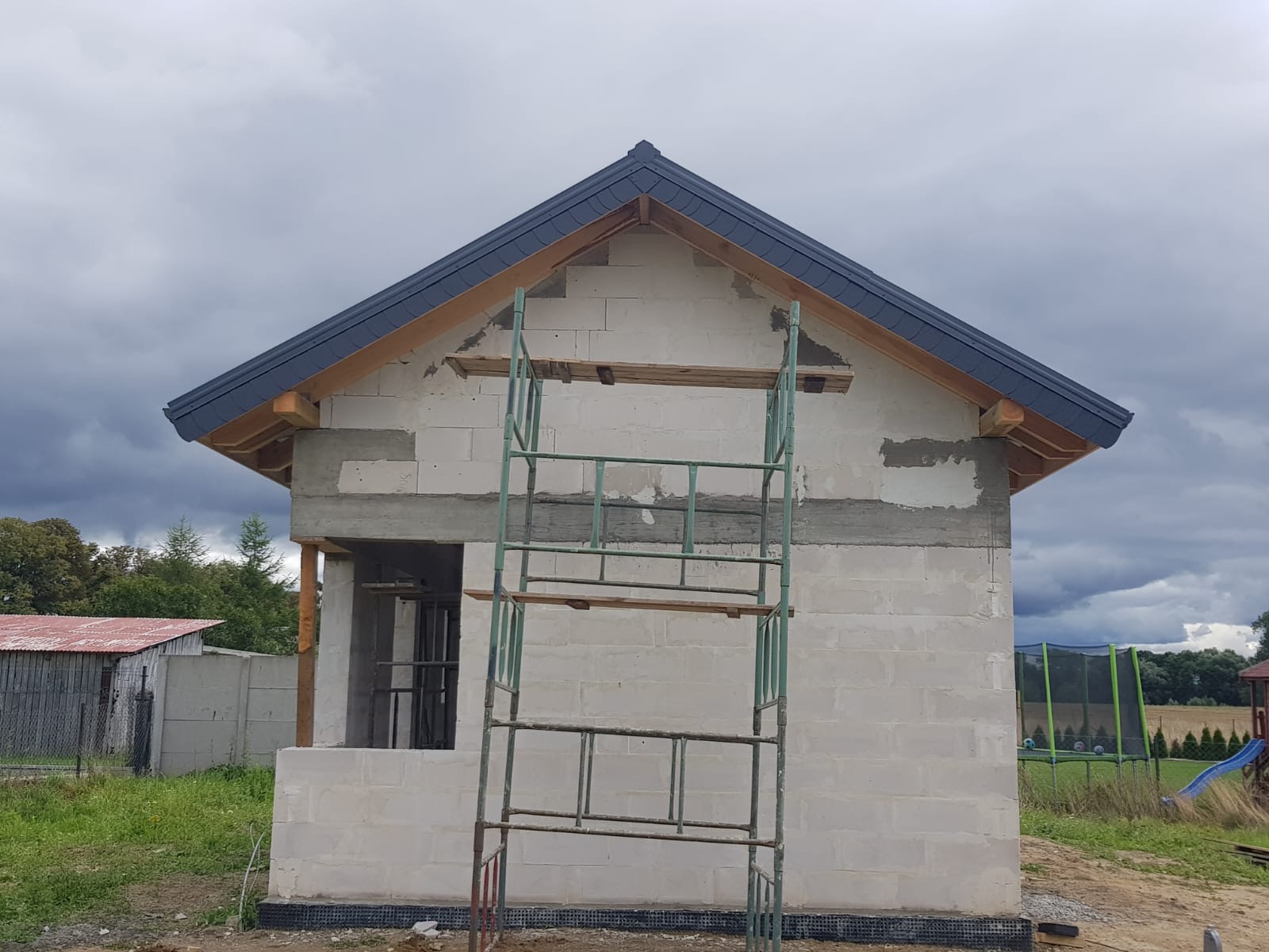 Surowy stan domu z białych bloczków, widoczne szare elementy konstrukcyjne, ciemna dachówka i metalowe rusztowanie na tle pochmurnego nieba.