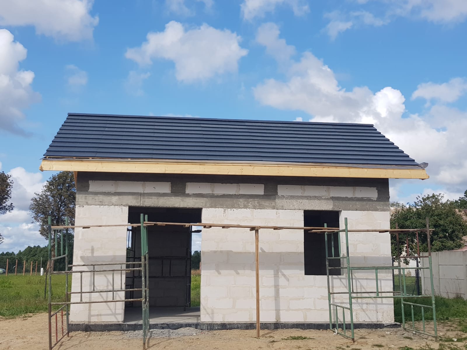 Niedokończony budynek z białych bloczków z zamontowanym ciemnym dachem, widoczne rusztowania i błękitne niebo z chmurami w tle.