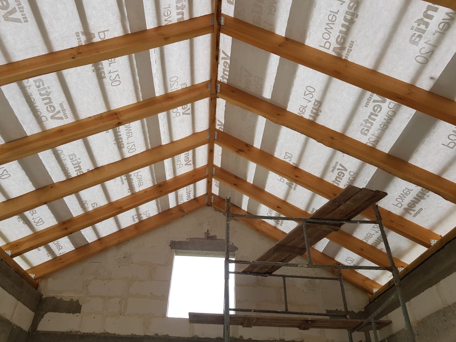 Konstrukcja dachu z drewnianych belek i membrany dachowej, widok z wnętrza budynku w trakcie budowy, z widocznym rusztowaniem i oknem.