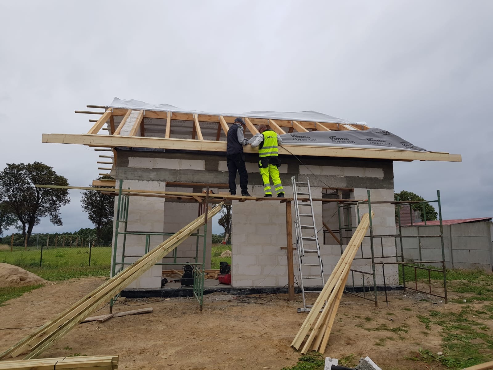 Budowa domu z bloczków: dwóch robotników montuje konstrukcję dachu na niewykończonym budynku, widoczne rusztowania i materiały budowlane.