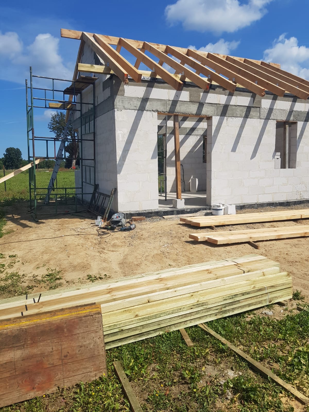 Surowy stan budowy domu z białych bloczków, widoczne drewniane elementy konstrukcji dachu, rusztowanie, narzędzia i stos drewna na trawie.