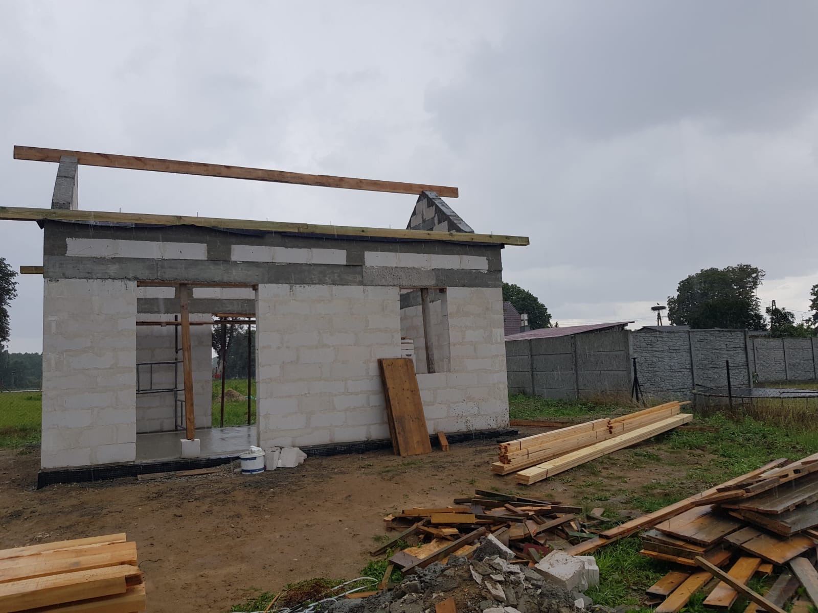 Surowy stan domu z betonu komórkowego z widocznymi belkami stropowymi i elementami szalunku, na ziemi leżą stosy drewna budowlanego, w tle betonowe ogrodzenie.