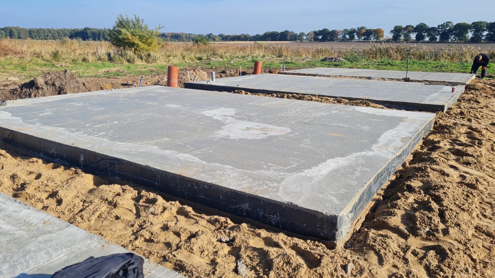 Wylane płyty fundamentowe zabezpieczone czarną izolacją, widoczne rury kanalizacyjne wystające z ziemi, piaszczyste podłoże i krajobraz polny w tle.