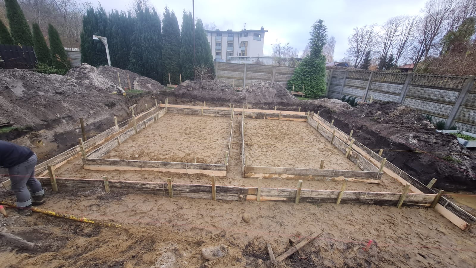 Przygotowanie terenu pod fundamenty: wykopane dwa prostokątne zagłębienia z drewnianym szalunkiem, widoczny pracownik w lewym dolnym rogu, ziemia wokół wykopu, w tle ogrodzenie i drzewa.