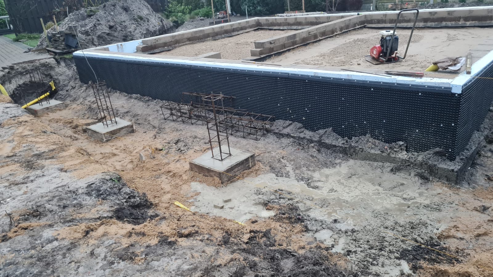 Etap budowy fundamentów: widoczne zbrojenia na betonowych podstawach, izolacja pionowa fundamentów z folii kubełkowej, częściowo zalany wykop, ubijarka w tle.