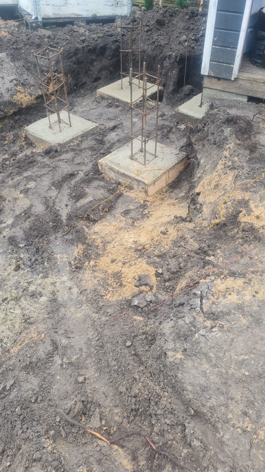 Wykopane fundamenty pod budynek z widocznymi betonowymi stopami i wystającymi zbrojeniami, otoczone mokrą ziemią i piaskiem.