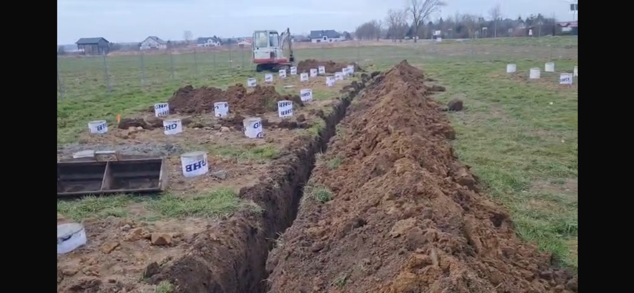 Wykop pod fundamenty z wyznaczonym obrysem, widoczny sprzęt budowlany i oznaczone punkty, teren budowy na zewnątrz.