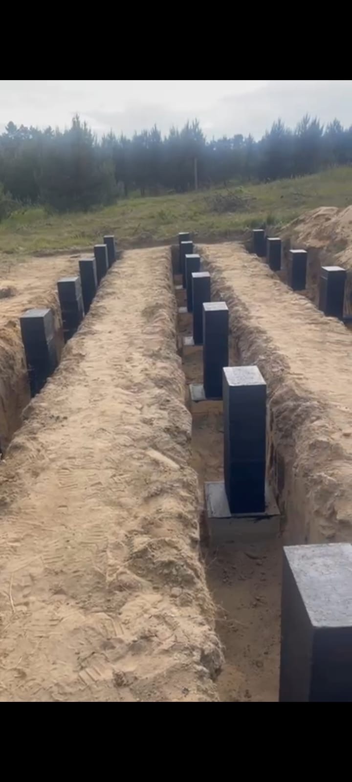 Przygotowane podłoże pod fundamenty z betonowymi słupami w wykopach, teren budowy na tle lasu. Widok z góry, proste linie wykopów.