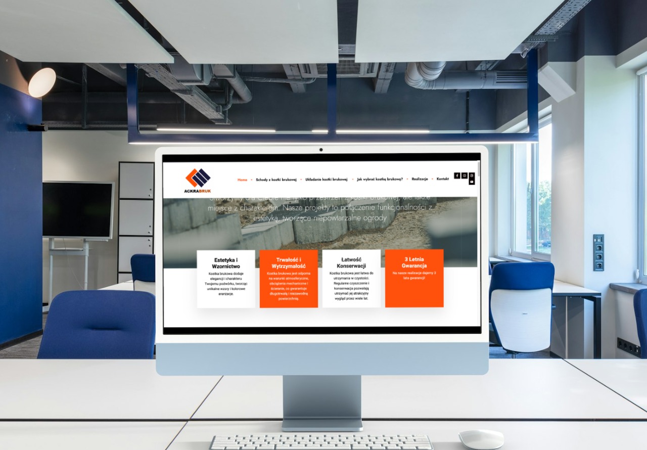 Monitor iMac wyświetlający stronę internetową firmy brukarskiej Ackrabruk, z widocznym logo i hasłami reklamowymi na tle nowoczesnego biura z niebieskimi akcentami.