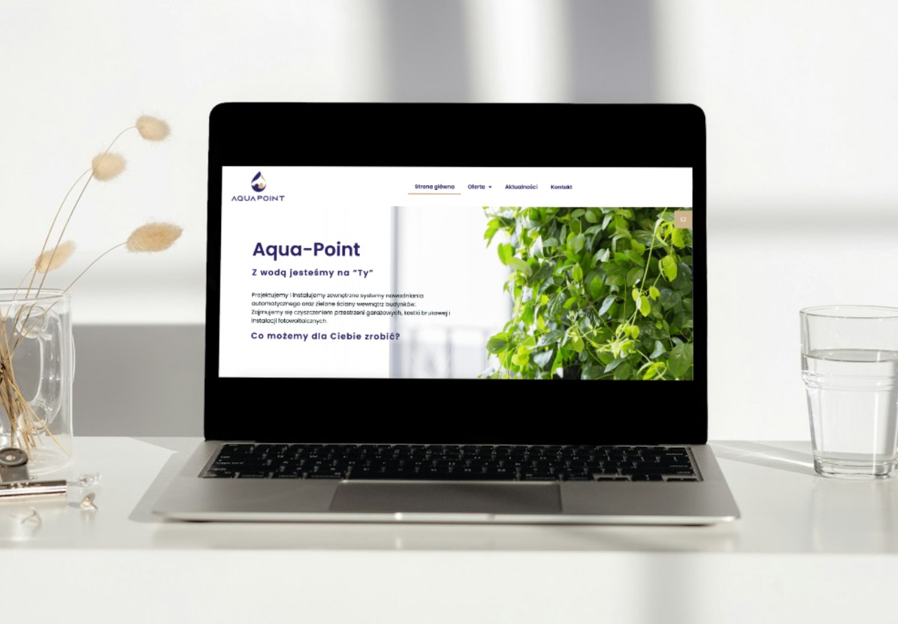 Laptop wyświetla stronę internetową firmy Aqua-Point z zieloną ścianą w tle, obok szklanka wody i dekoracyjny wazonik z suszem, na jasnym biurku.