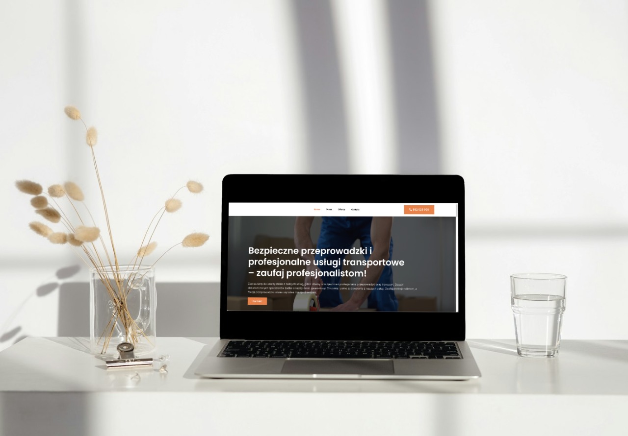 Laptop wyświetlający stronę internetową firmy transportowej, ustawiony na białym biurku obok szklanki wody i wazonu z suszonymi kwiatami.