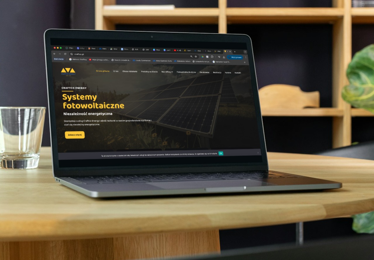 Laptop wyświetla stronę internetową firmy zajmującej się systemami fotowoltaicznymi, obok stoi szklanka na drewnianym stole.