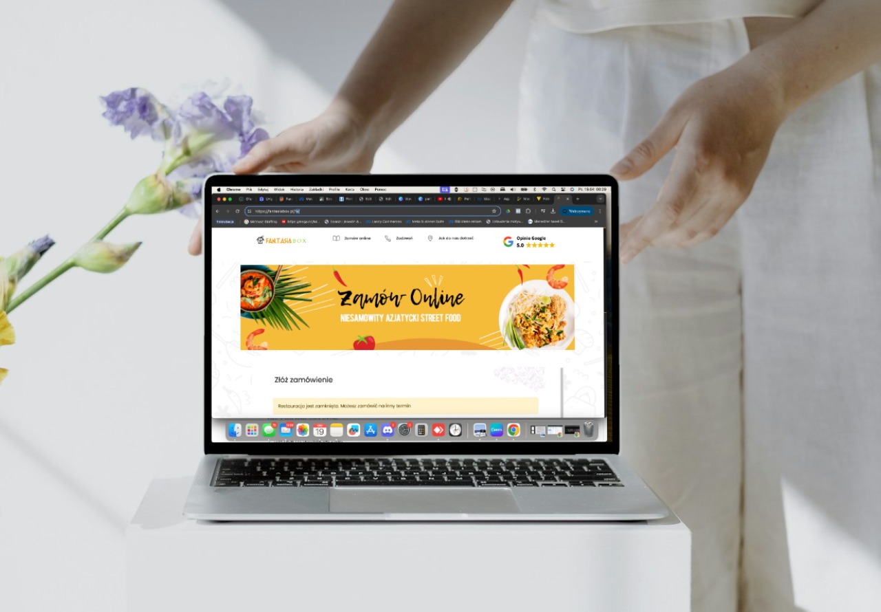 Kobieta w białym ubraniu prezentuje na laptopie stronę internetową restauracji azjatyckiej z możliwością zamówień online, obok laptopa kompozycja kwiatowa.