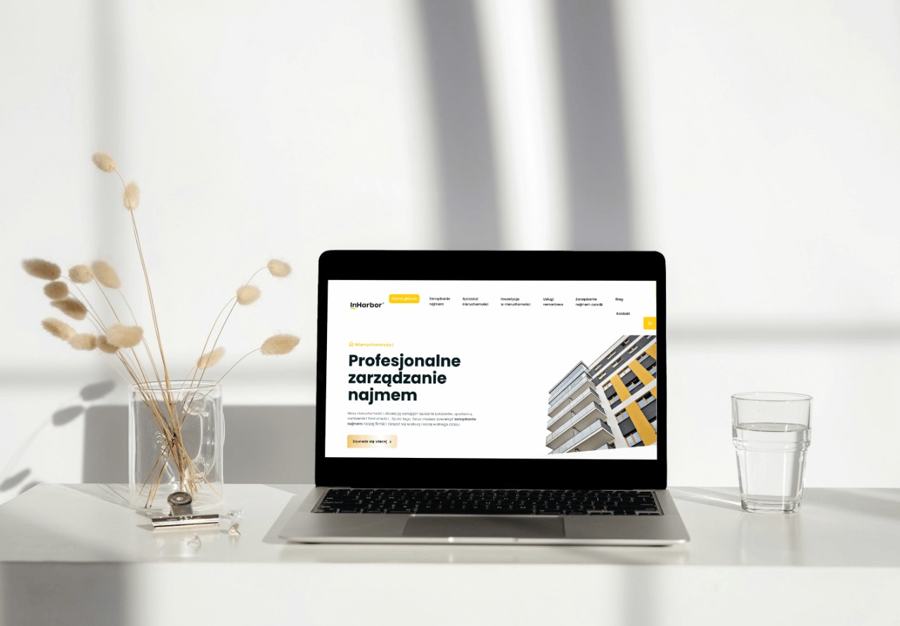 Laptop wyświetla stronę internetową firmy InHarbor z branży nieruchomości, obok szklanka wody i dekoracyjne trawy w wazonie, minimalistyczne biuro z białym tłem.
