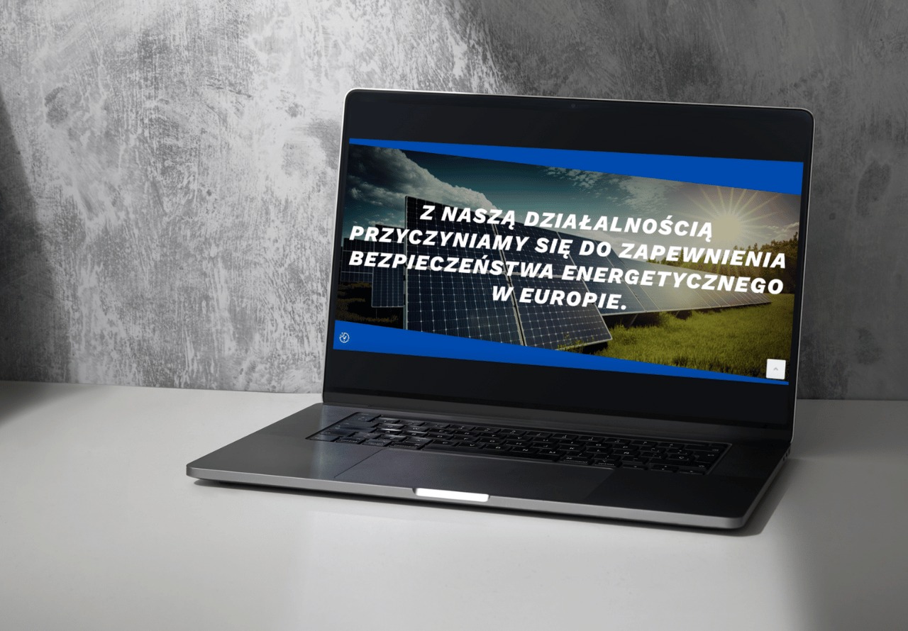 Ekran laptopa wyświetla stronę internetową z panelami słonecznymi na tle słonecznego nieba i hasłem o bezpieczeństwie energetycznym Europy, ustawiony na białym biurku przed szarą ścianą.