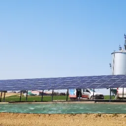 Długi rząd paneli słonecznych zamontowanych na konstrukcji w polu, z widocznymi silosami zbożowymi w tle i dwoma samochodami serwisowymi parkującymi obok instalacji.