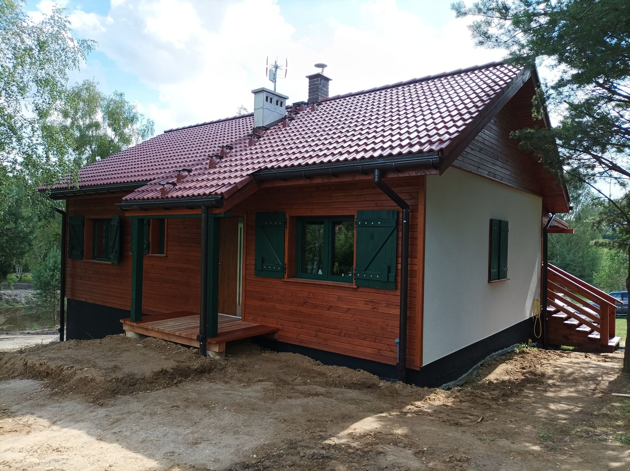 Gotowy drewniany dom z czerwonym dachem i zielonymi okiennicami w Stawigudzie, widoczne elementy wykończeniowe i zagospodarowanie terenu.