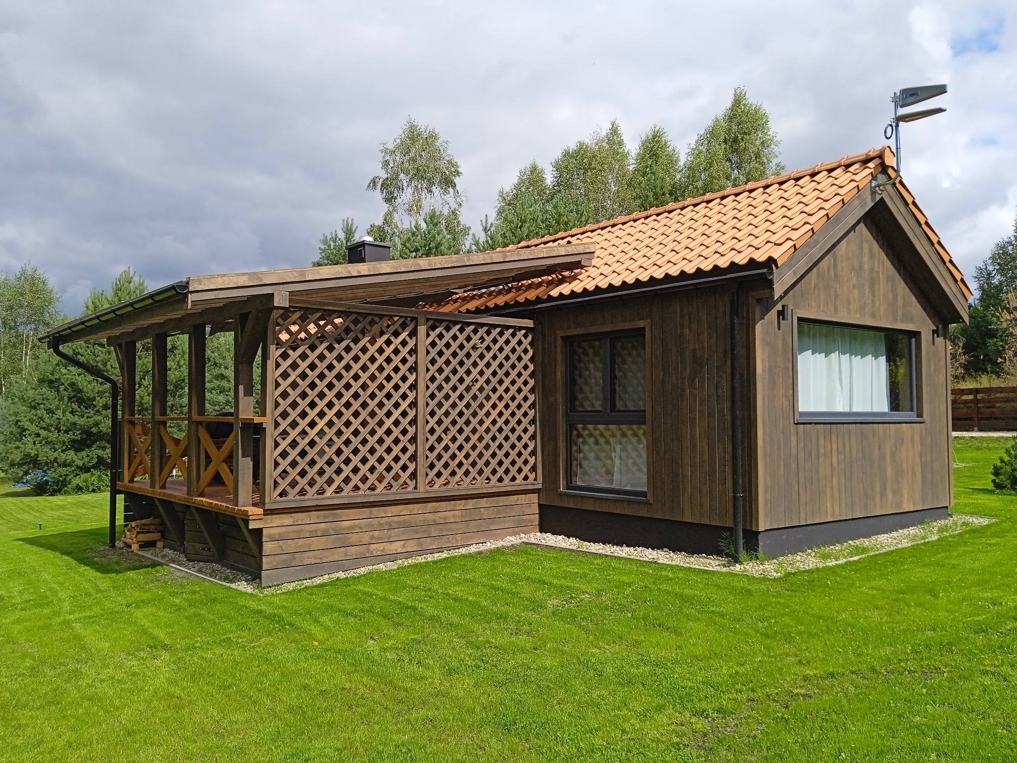 Nowoczesny dom z ciemnego drewna z zadaszonym tarasem z kratownicą, pomarańczowa dachówka i równo przystrzyżony trawnik w słoneczny dzień.