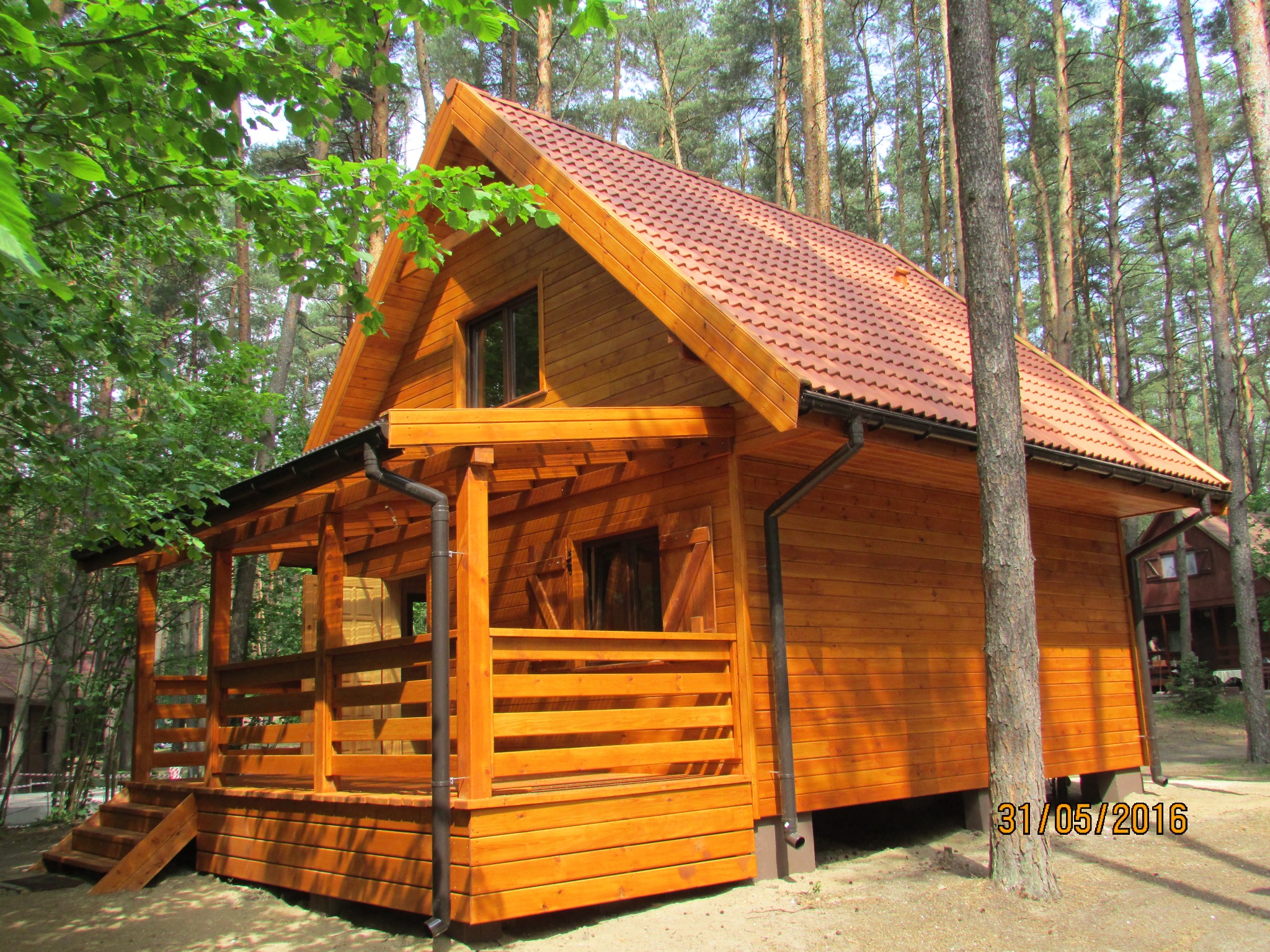 Nowoczesny, drewniany dom szkieletowy z zadaszonym tarasem, wykończony czerwoną dachówką, otoczony sosnowym lasem w słoneczny dzień.