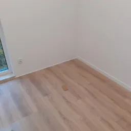 Jasne panele podłogowe w nowym pokoju, widok z góry na fragment podłogi, listwy przypodłogowe i białe ściany, w tle okno z widokiem na drzewa.