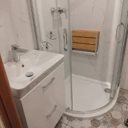 Nowoczesna łazienka po remoncie: biała umywalka z szafką, kabina prysznicowa z siedziskiem z drewna, mozaika na podłodze, widoczny fragment butelki z płynem do czyszczenia.