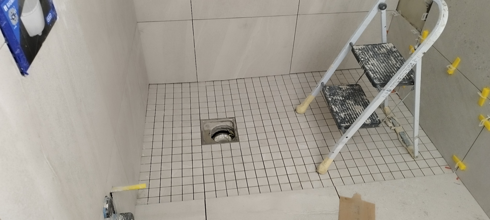 Ukończona posadzka z płytek w kabinie prysznicowej, widoczny odpływ liniowy, drabina malarska i żółte krzyżyki dystansowe na ścianie.