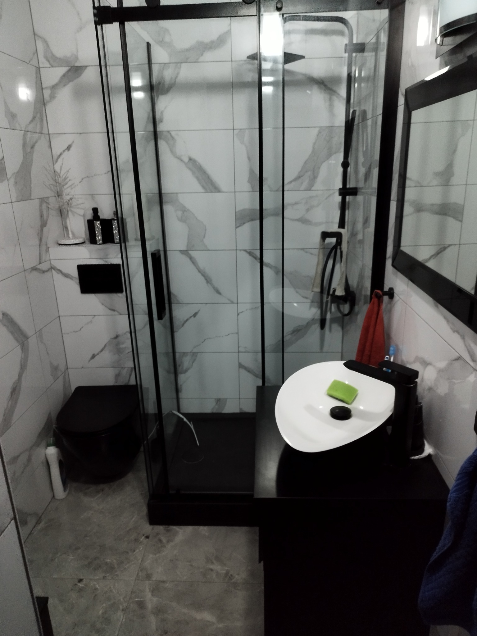 Nowoczesna łazienka z czarnymi akcentami: kabina prysznicowa typu walk-in z czarnymi ramami, wisząca toaleta i umywalka nablatowa na czarnej szafce, płytki imitujące marmur na ścianach i podłodze.