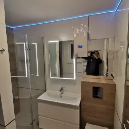 Nowoczesna łazienka po remoncie z kabiną prysznicową, umywalką z szafką, toaletą typu Geberit i podświetleniem LED podsufitowym oraz lustra, uchwycona z perspektywy osoby robiącej zdjęcie w lustrze.