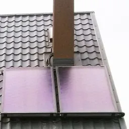 Dwa płaskie panele słoneczne zamontowane na dachu pokrytym szarą dachówką, obok okna dachowego i komina. Widoczne metalowe ramy i rury. Ujęcie z dołu.