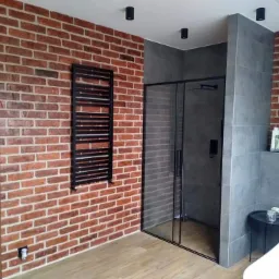 Nowoczesna łazienka z czerwoną ścianą z cegły, czarną kabiną prysznicową i grzejnikiem drabinkowym. Widoczna drewnopodobna podłoga i szare płytki w strefie prysznica. Minimalistyczne oświetlenie...