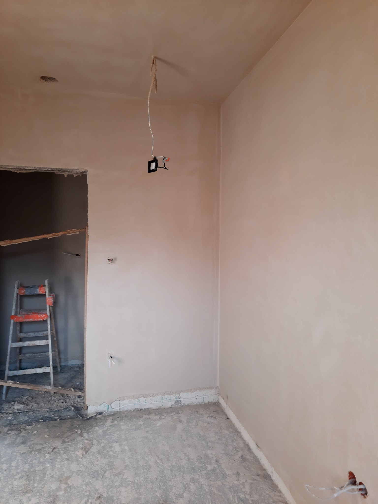 Pokój w trakcie prac wykończeniowych: świeżo otynkowane ściany w jasnym odcieniu, widoczne wystające przewody elektryczne, drabina oparta o framugę drzwiową, surowy beton na podłodze.