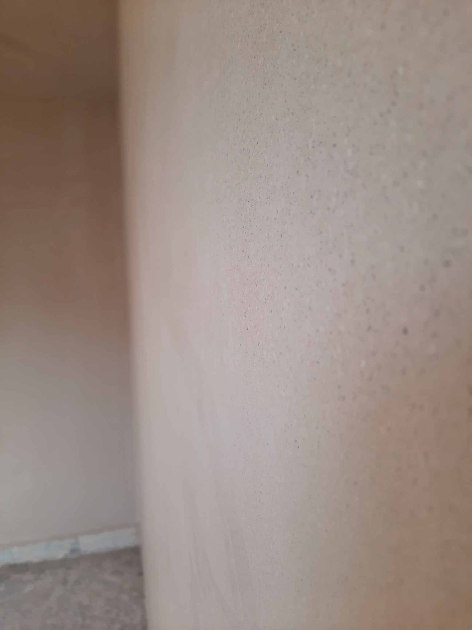 Świeżo nałożony, jasny tynk na ścianie wewnętrznej, widoczne drobne nierówności i faktura, narożnik ściany w tle, słabe oświetlenie.