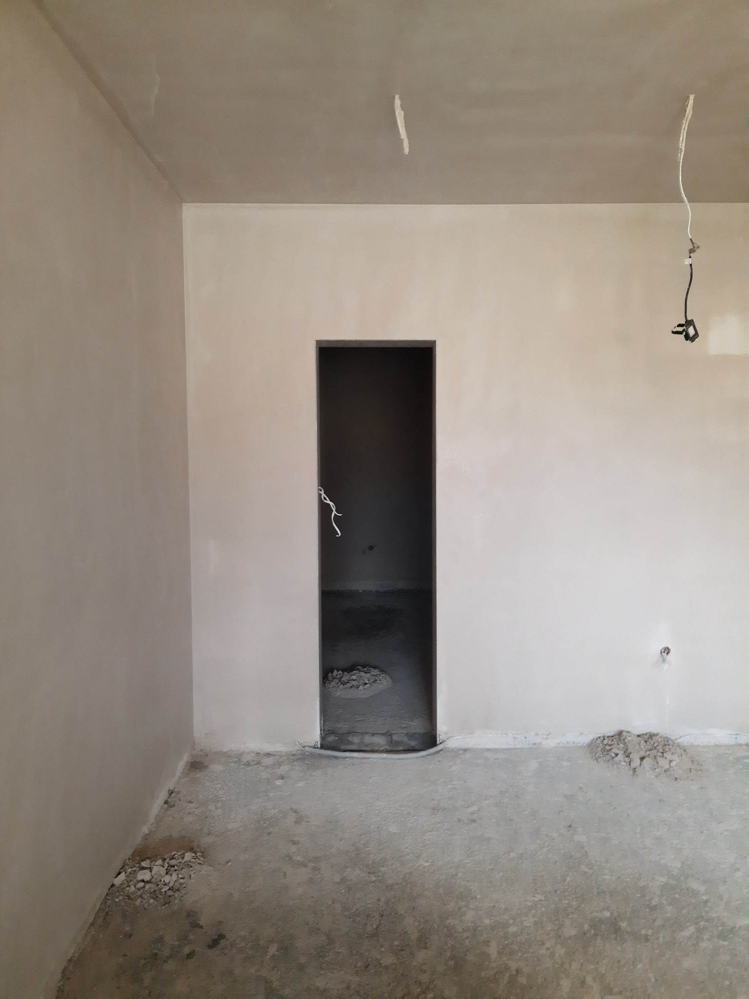 Wnętrze w trakcie prac wykończeniowych: ściany i sufit pokryte świeżym, jasnym tynkiem, widoczne przewody elektryczne zwisające z sufitu, resztki materiałów budowlanych na podłodze, surowy otwór...